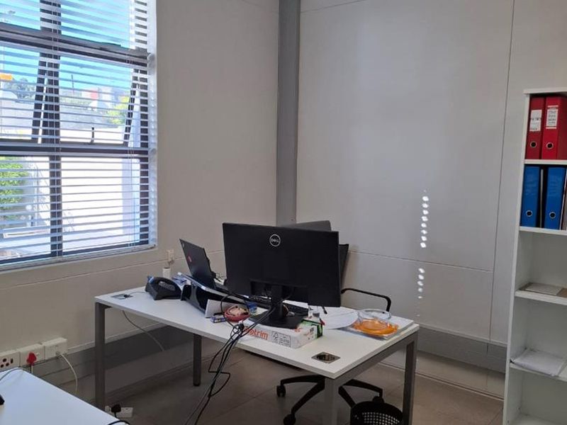 A functional office space featuring tiled flooring, a wall-mounted air conditioning unit, and large windows with venetian blinds for natural light control.