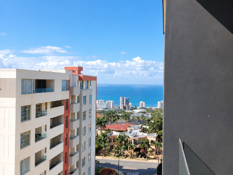 Elevated view from a balcony overlooking the street, surrounding residential buildings, and the ocean in the distance.