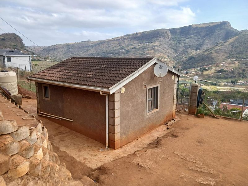 Rear view of a detached house featuring a tiled roof, satellite dish, and stone retaining wall, set against a mountainous landscape.