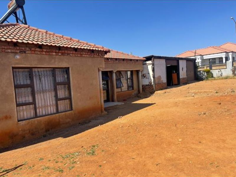 Front exterior view of a single-story house featuring a tiled roof, solar water heater, and an attached garage with an unpaved yard area.