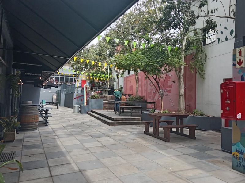 An outdoor paved courtyard area featuring raised timber decking, built-in planters, and a retractable awning, suitable for communal office use or break-out space.