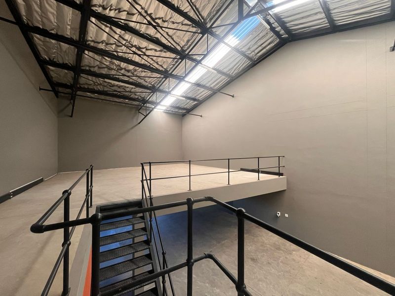 Spacious industrial utility area featuring a mezzanine level, metal staircase with safety railings, exposed steel roof trusses, and natural light from skylight panels.