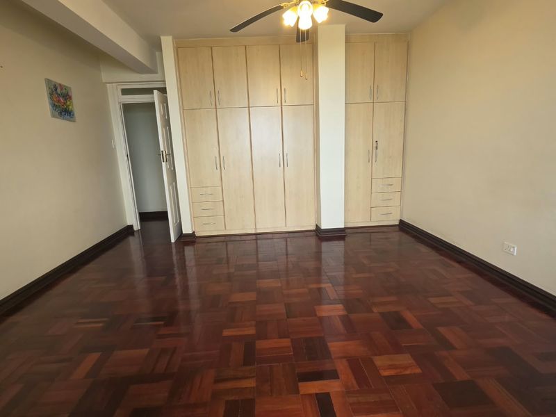 Spacious bedroom featuring polished parquet flooring, extensive built-in wooden cupboards, and a ceiling fan, offering a clean and functional living space.