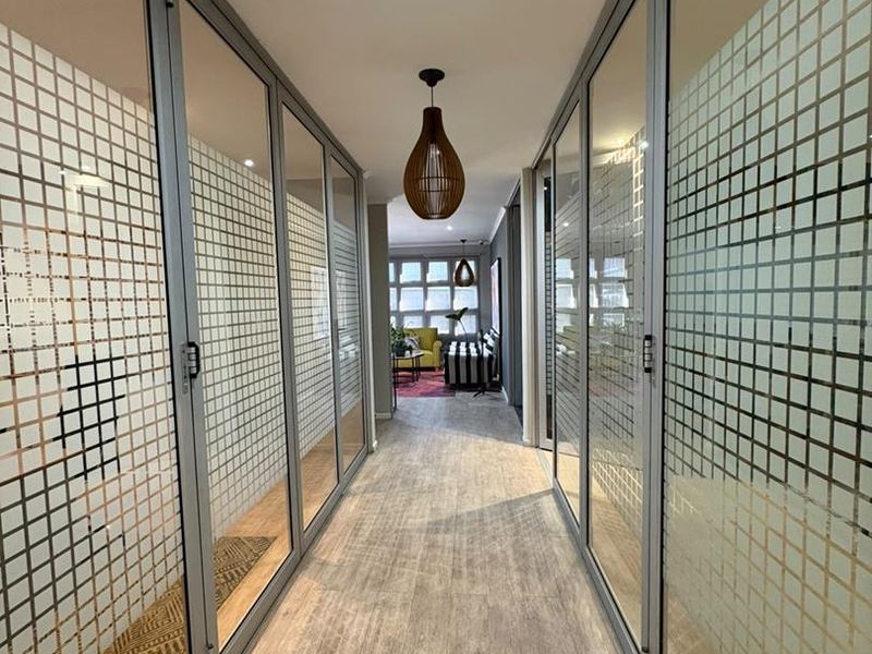 A modern office hallway featuring glass partition walls with privacy film and wood-look laminate flooring, leading to an open-plan workspace.