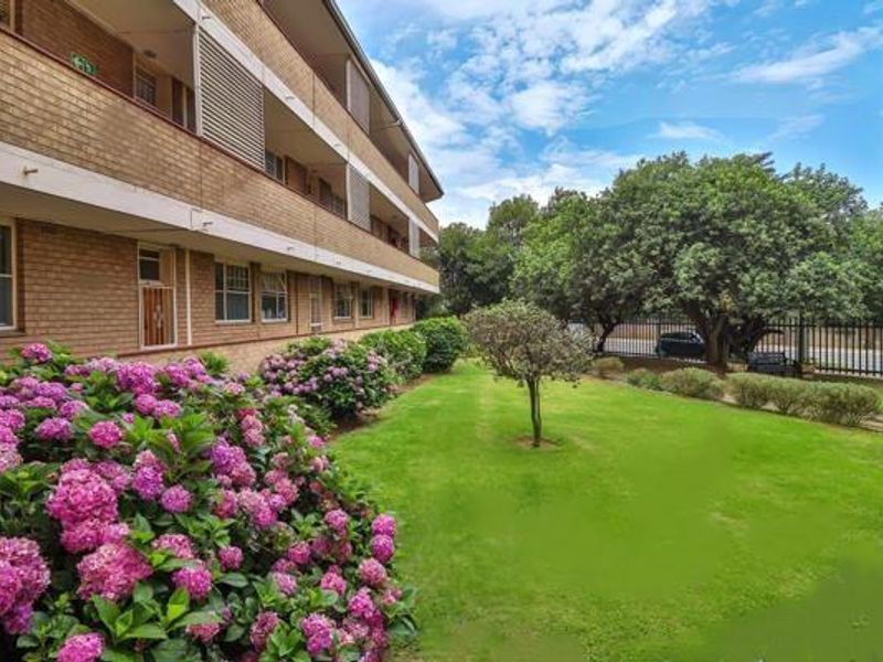 A view of the apartment building's rear facade featuring brick construction, multiple balconies, and a well-maintained communal garden area with mature trees.