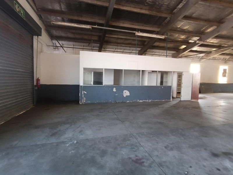 Spacious industrial utility area featuring high ceilings with exposed steel trusses, a large roller shutter door, and a partitioned office enclosure.