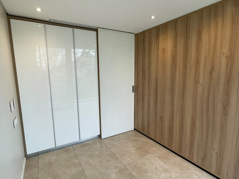 A modern, empty bedroom featuring sleek built-in cupboards with white gloss and wood-grain finishes, complemented by neutral tiled flooring and recessed ceiling lighting.