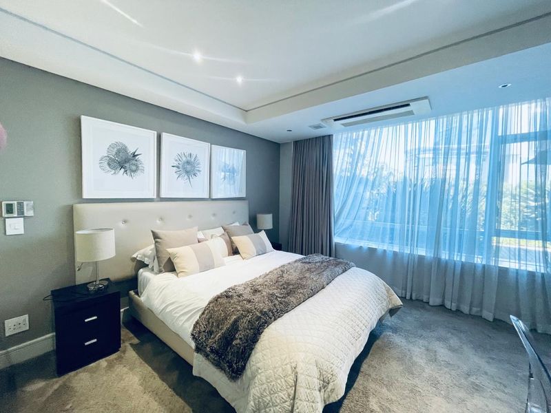 A well-appointed bedroom featuring recessed ceiling lighting, integrated ducted air conditioning, and large windows with sheer curtains, finished with neutral carpeting.