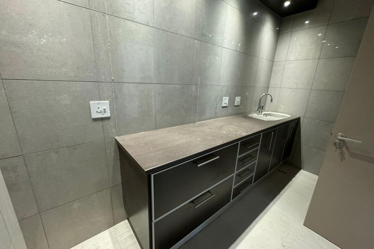 A modern kitchenette area featuring dark cabinetry, stone-look countertops, a stainless steel sink, and full-height grey wall tiling.