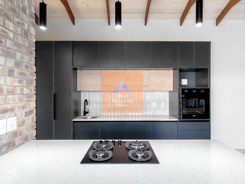 Modern kitchen featuring sleek dark cabinetry, stone countertops, a gas hob, exposed brickwork, and timber ceiling beams in a contemporary open-plan layout.