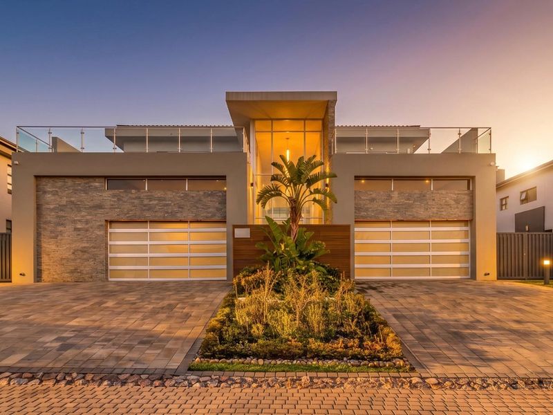 A modern double-story residence featuring a symmetrical facade, stone cladding, dual garage doors, and a glass-railed upper balcony at dusk.
