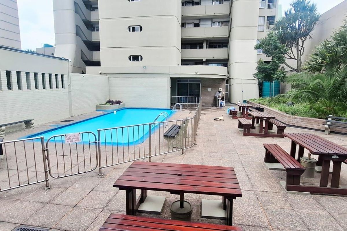 Communal outdoor pool area featuring a paved terrace, metal safety fencing, and landscaped garden beds within an apartment complex setting.