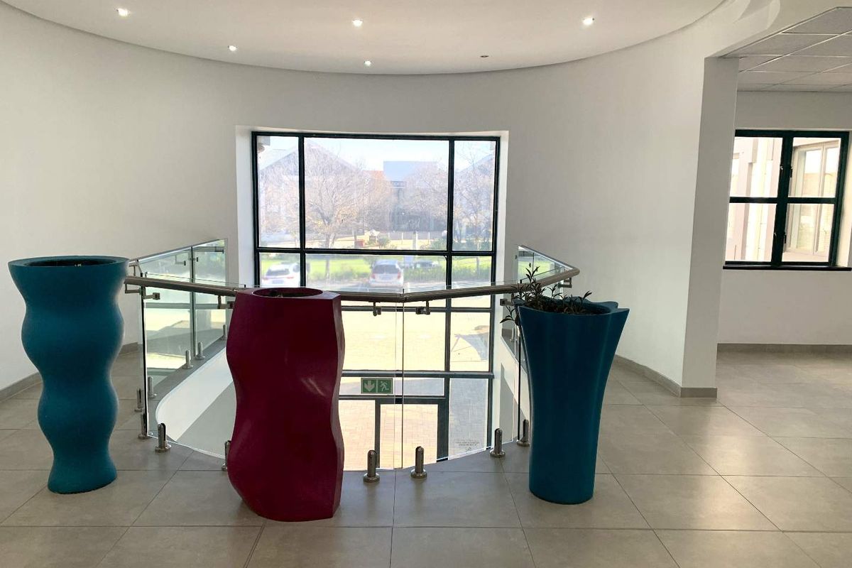 A bright and spacious office hallway featuring modern tiled flooring, a glass balustrade overlooking the entrance, and large windows providing ample natural light.