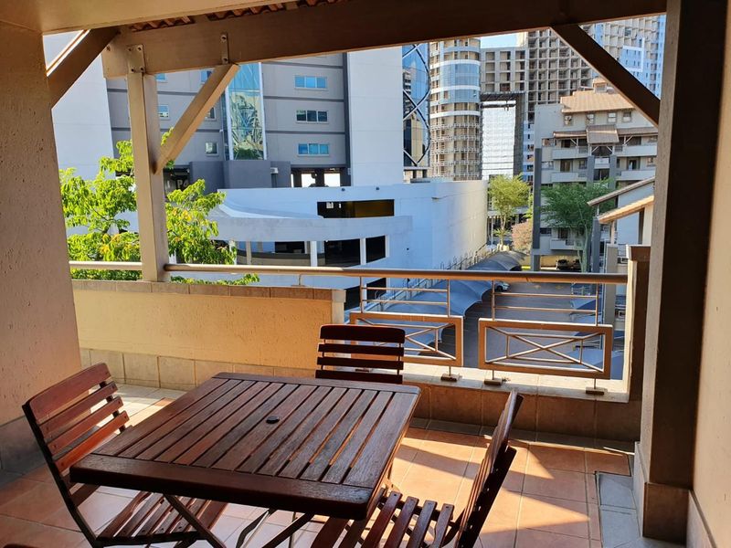 A covered balcony featuring tiled flooring, a metal safety railing, and timber structural beams, offering an urban view of the surrounding apartment complex.