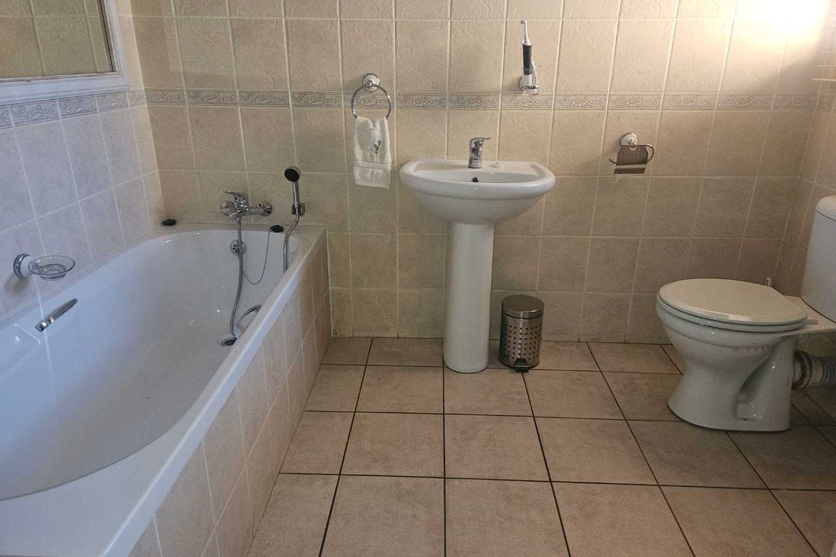 A functional bathroom featuring a built-in bathtub with shower mixer, pedestal basin, and toilet, finished with neutral wall and floor tiling.