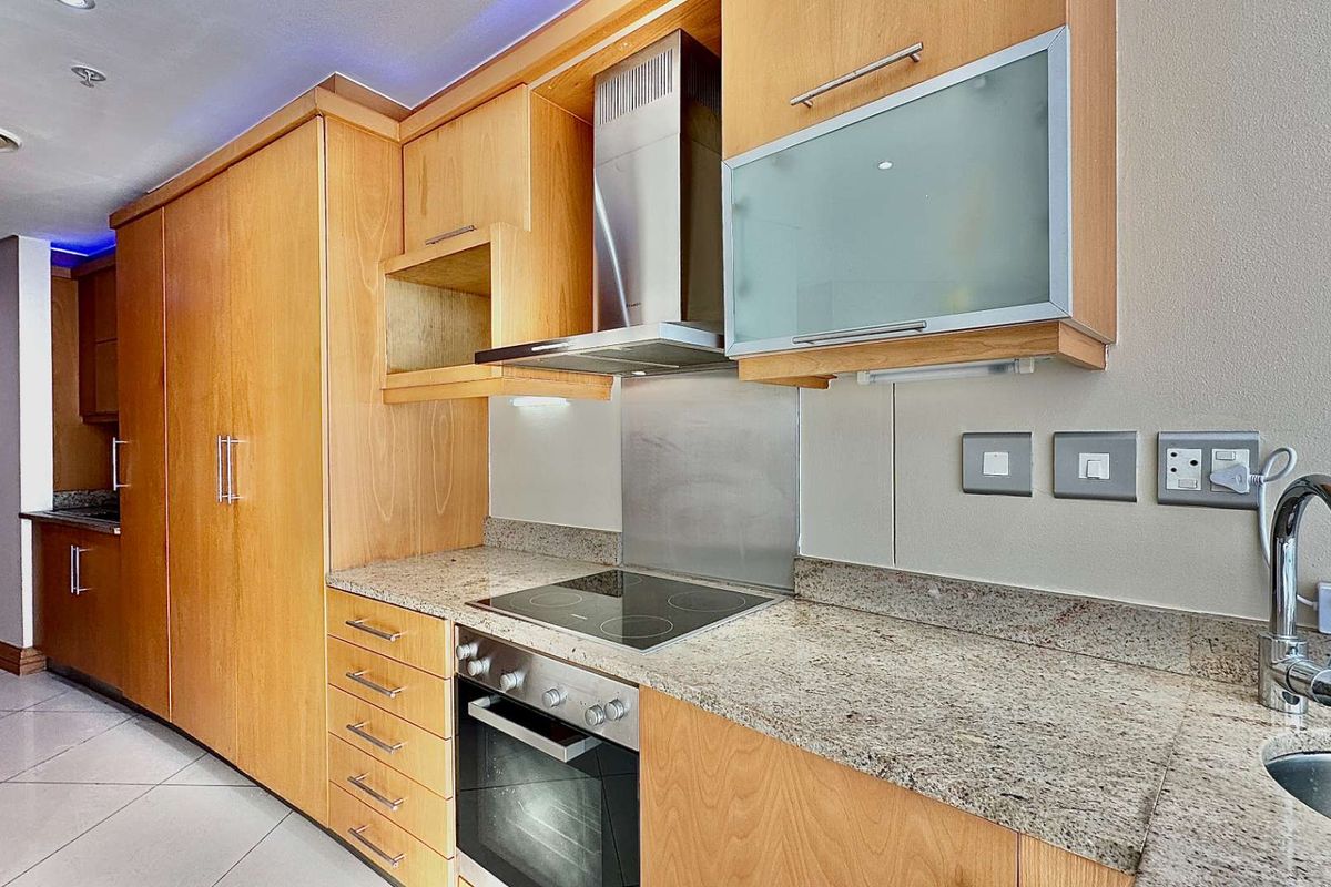 A modern kitchen featuring light wood cabinetry, granite countertops, a built-in oven, electric hob, stainless steel extractor fan, and a stainless steel sink.