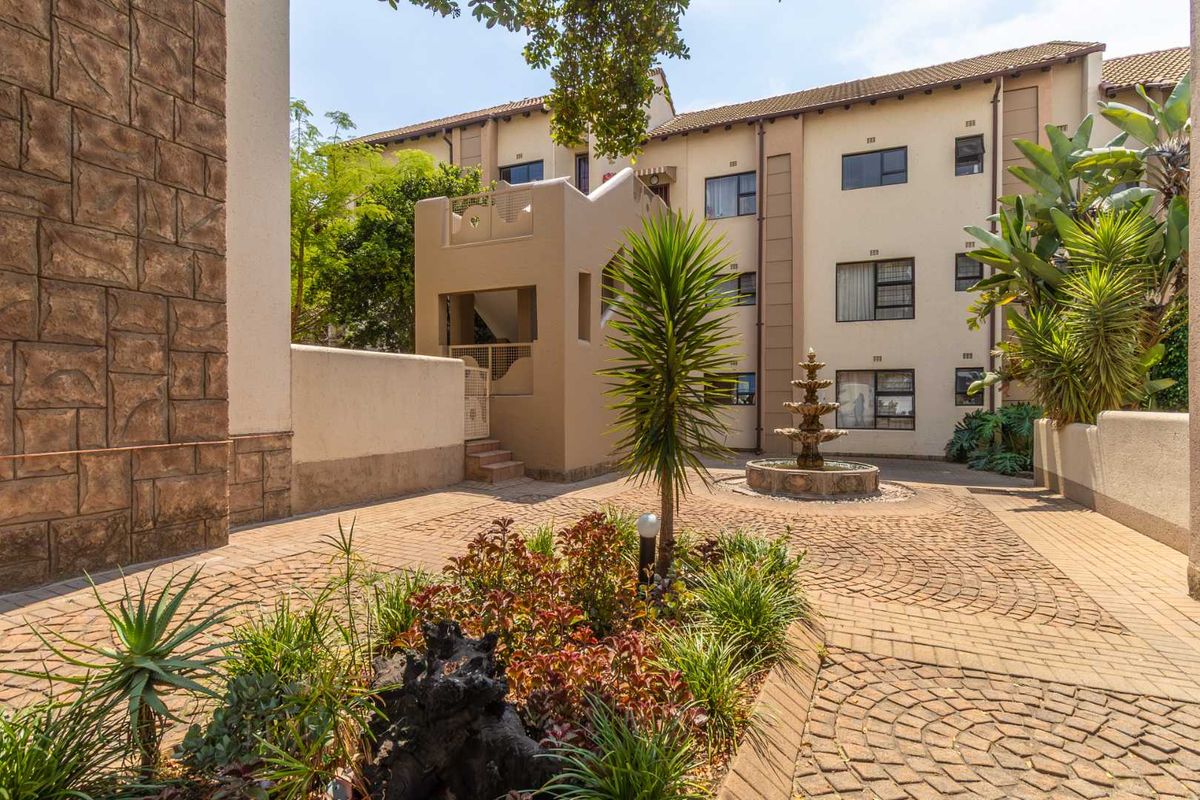 A well-maintained apartment complex courtyard featuring paved walkways, a central water fountain, stone-clad architectural details, and established garden landscaping.