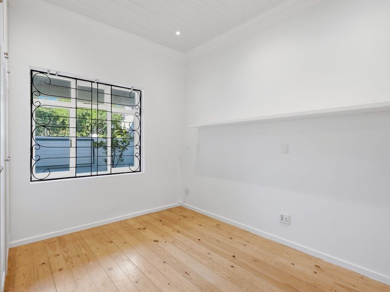A bright, empty bedroom featuring polished timber flooring, a tongue and groove ceiling, built-in cupboards, and a window with decorative security bars.
