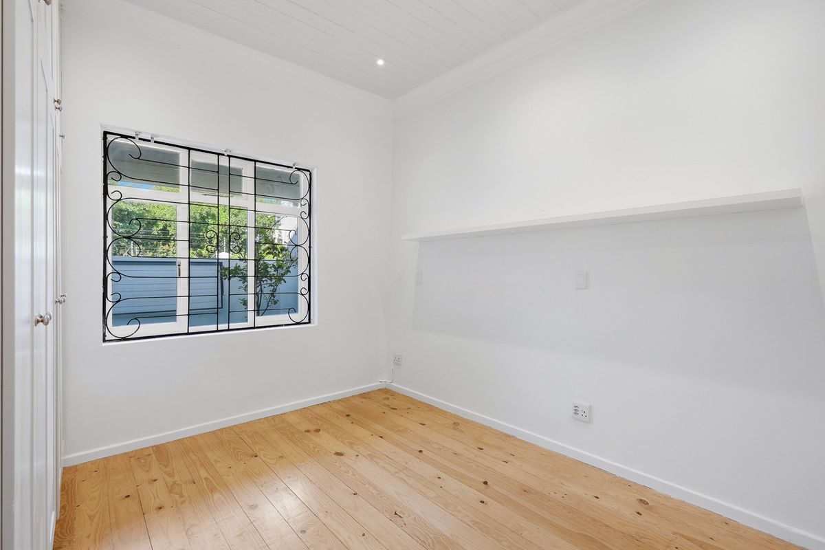 A bright, empty bedroom featuring polished timber flooring, a tongue and groove ceiling, built-in cupboards, and a window with decorative security bars.