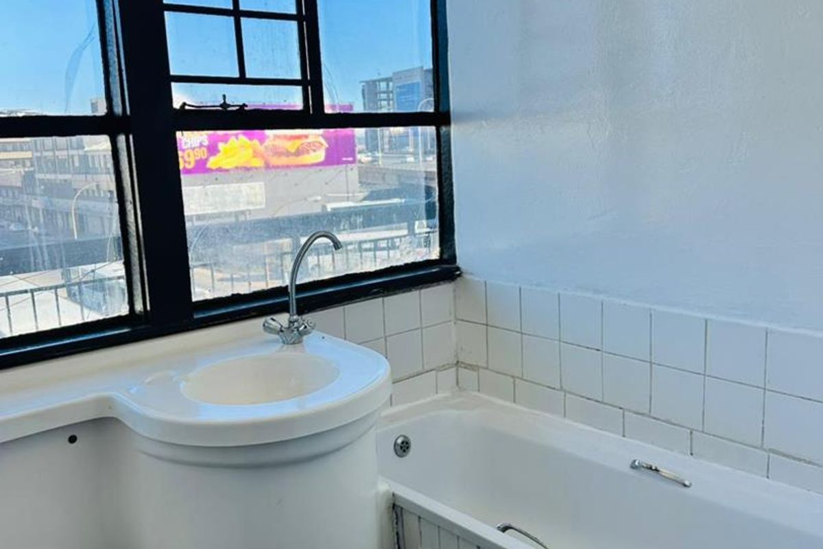 A compact bathroom featuring a bathtub, basin, and toilet with a large metal-framed window, requiring maintenance and cosmetic updates to the fixtures and finishes.