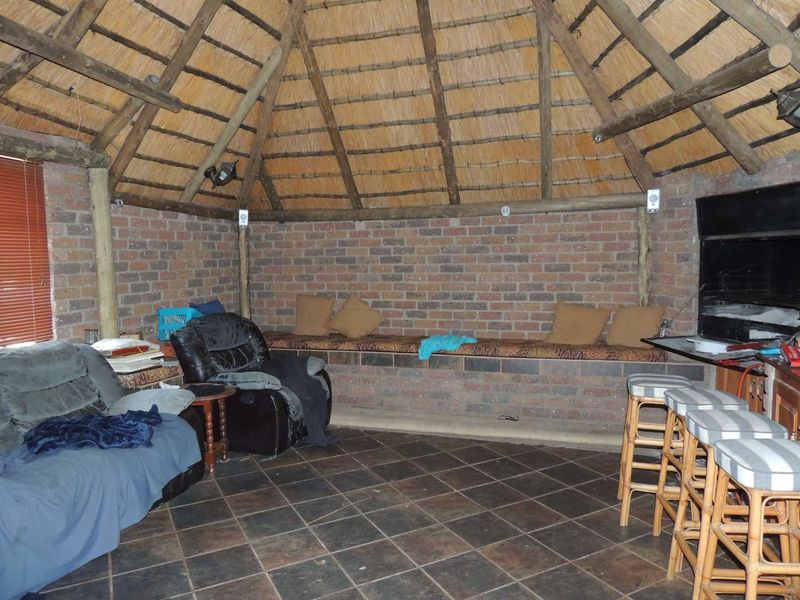 Rustic covered patio area featuring a traditional thatched roof, exposed brick walls, tiled flooring, and a built-in braai for outdoor entertaining.