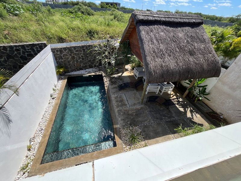 An elevated view of a private swimming pool area featuring a thatched gazebo, stone boundary wall, and paved patio space.
