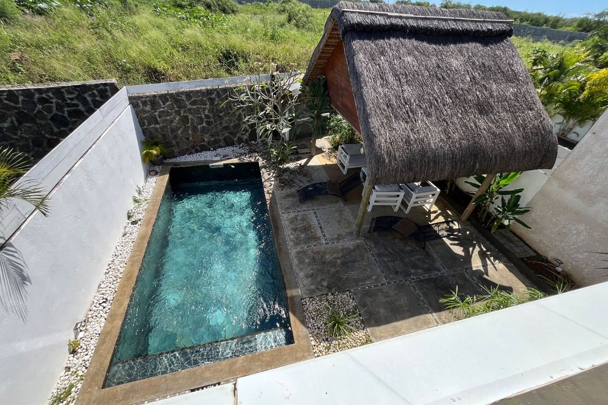 An elevated view of a private swimming pool area featuring a thatched gazebo, stone boundary wall, and paved patio space.