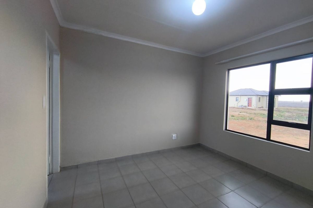A clean, empty bedroom featuring neutral wall paint, tiled flooring, and a large window providing natural light and a view of the surrounding area.
