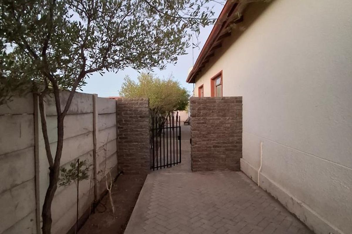 A side pathway featuring paved brick flooring, a precast concrete boundary wall, and a gated entrance leading to the rear of the property.