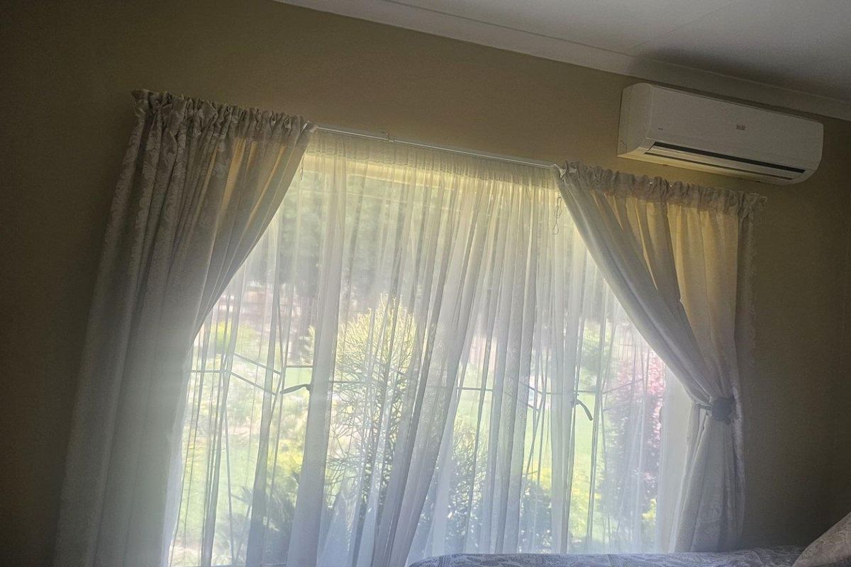 A bedroom featuring a wall-mounted air conditioning unit and a large window with curtains overlooking the garden.