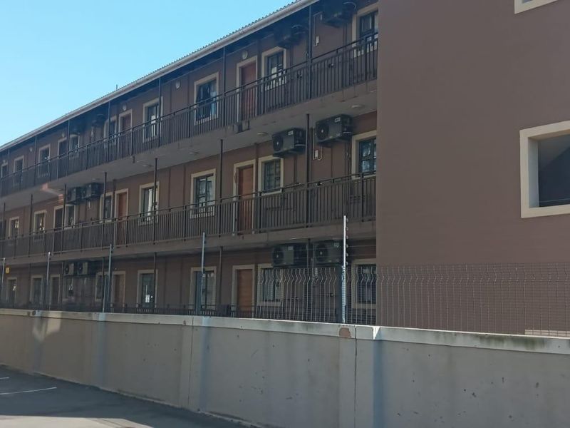 Exterior view of a multi-story apartment complex featuring external air conditioning units, metal balcony railings, and a secure concrete boundary wall.