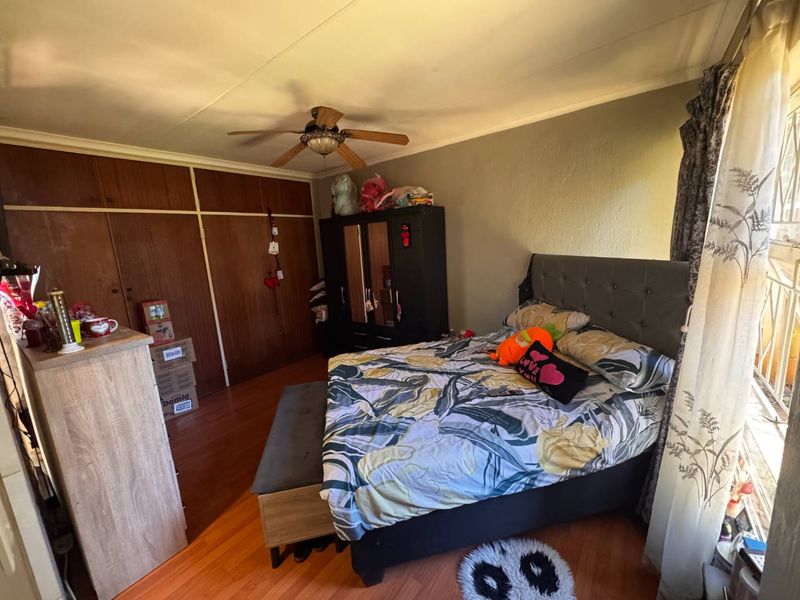 A bedroom featuring laminate flooring, built-in wooden cupboards, and a ceiling fan, with security bars installed on the window.