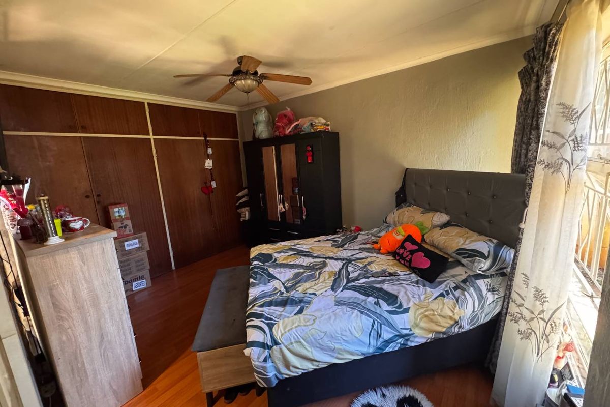 A bedroom featuring laminate flooring, built-in wooden cupboards, and a ceiling fan, with security bars installed on the window.