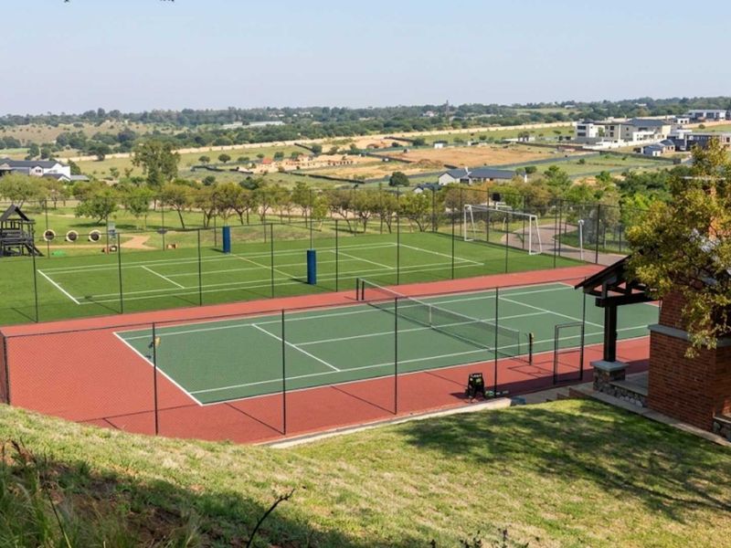 Well-maintained sports facility featuring a tennis court, soccer area, and a brick pavilion with an outdoor fireplace, set within a landscaped estate.