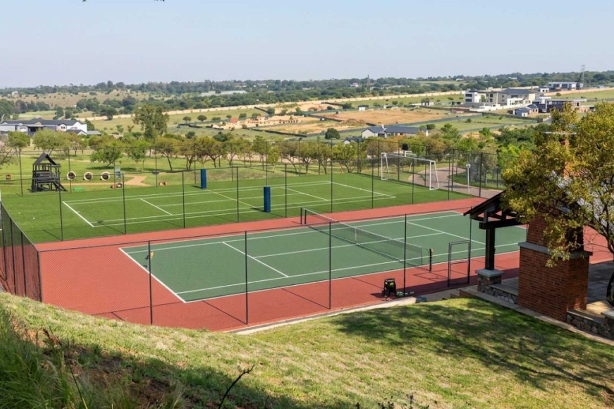 Well-maintained sports facility featuring a tennis court, soccer area, and a brick pavilion with an outdoor fireplace, set within a landscaped estate.