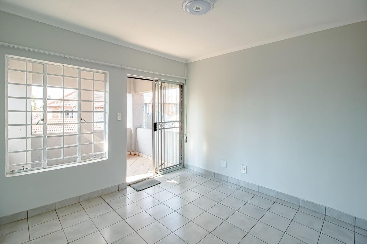 A bright, empty living area featuring neutral tiled flooring, a large window with security bars, and a sliding door leading to a private balcony.