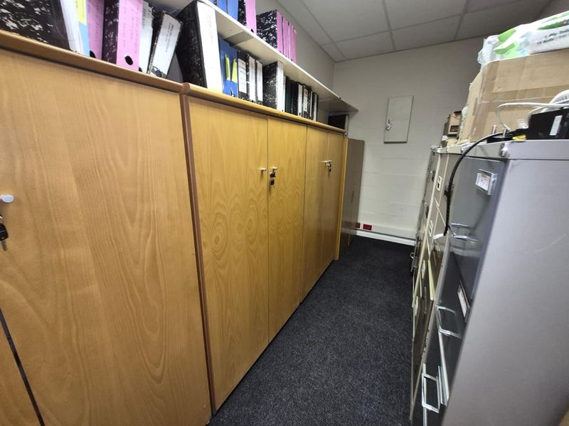 A functional office storage area featuring extensive built-in wooden cabinetry, overhead shelving, and durable carpet flooring.