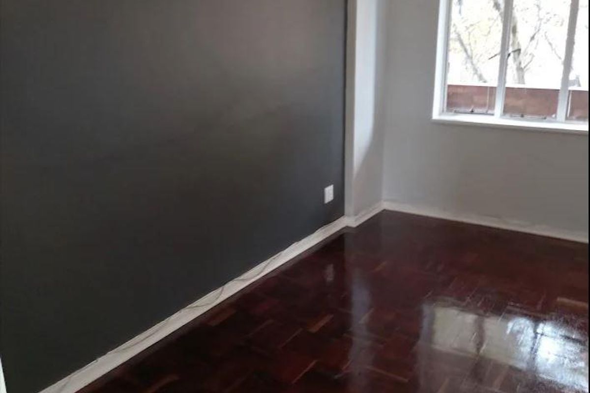 A bright, empty bedroom featuring polished dark parquet flooring and a large multi-pane window with a feature wall.