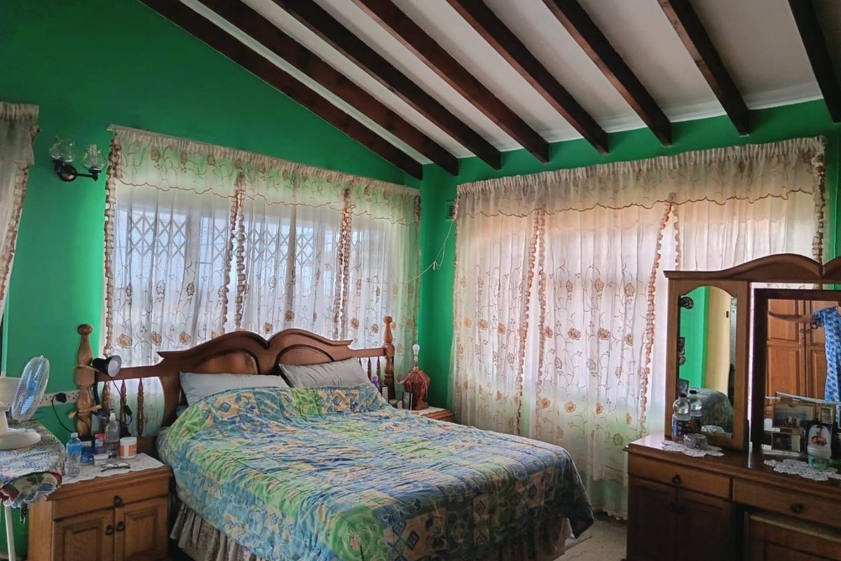 A bedroom featuring exposed timber ceiling beams and large windows with security gates, offering a dated aesthetic ready for modernization.