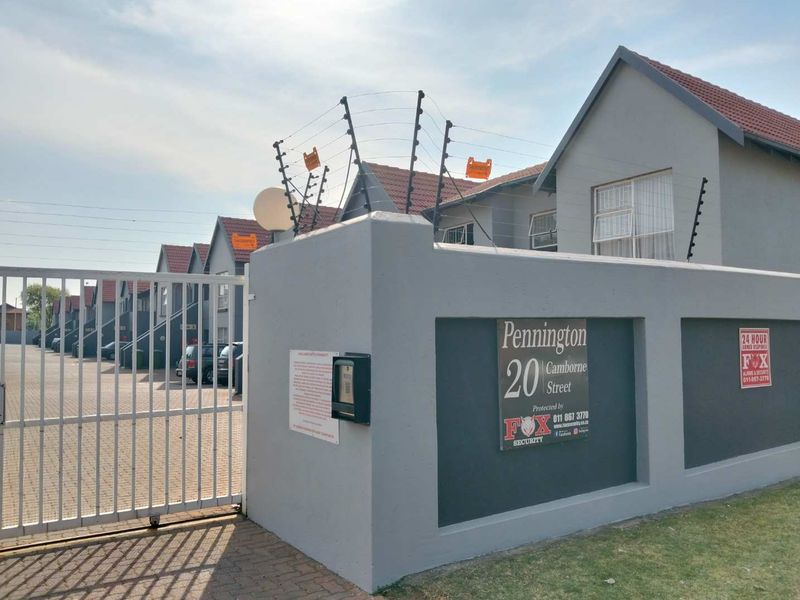Front entrance view of the townhouse complex featuring a secure gated access point, perimeter wall with electric fencing, and paved driveway.