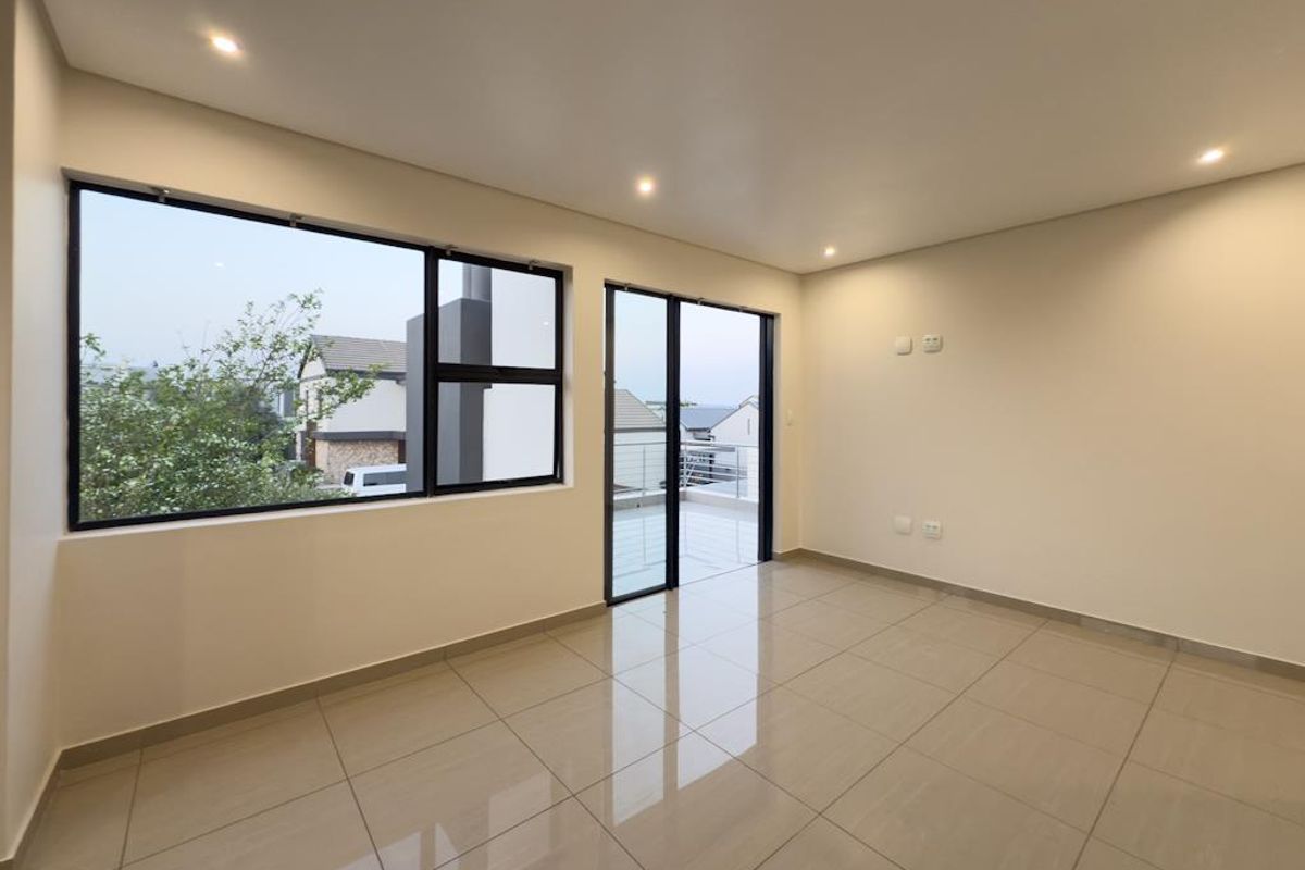 A bright, modern living area featuring large windows, glass sliding doors leading to a balcony, recessed ceiling lighting, and polished floor tiles.