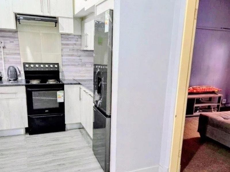 Compact kitchen featuring built-in cabinetry, a modern stove with extractor fan, and tiled backsplash, with an adjacent view into a bedroom area.