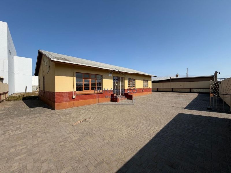 Rear exterior view of a single-story building featuring a paved courtyard, brick and plaster facade, and secure gated entrance.