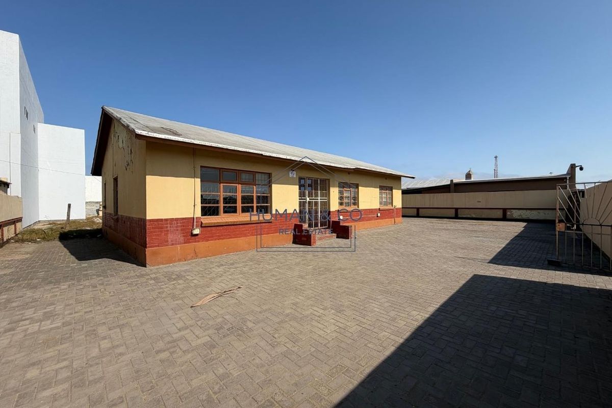 Rear exterior view of a single-story building featuring a paved courtyard, brick and plaster facade, and secure gated entrance.