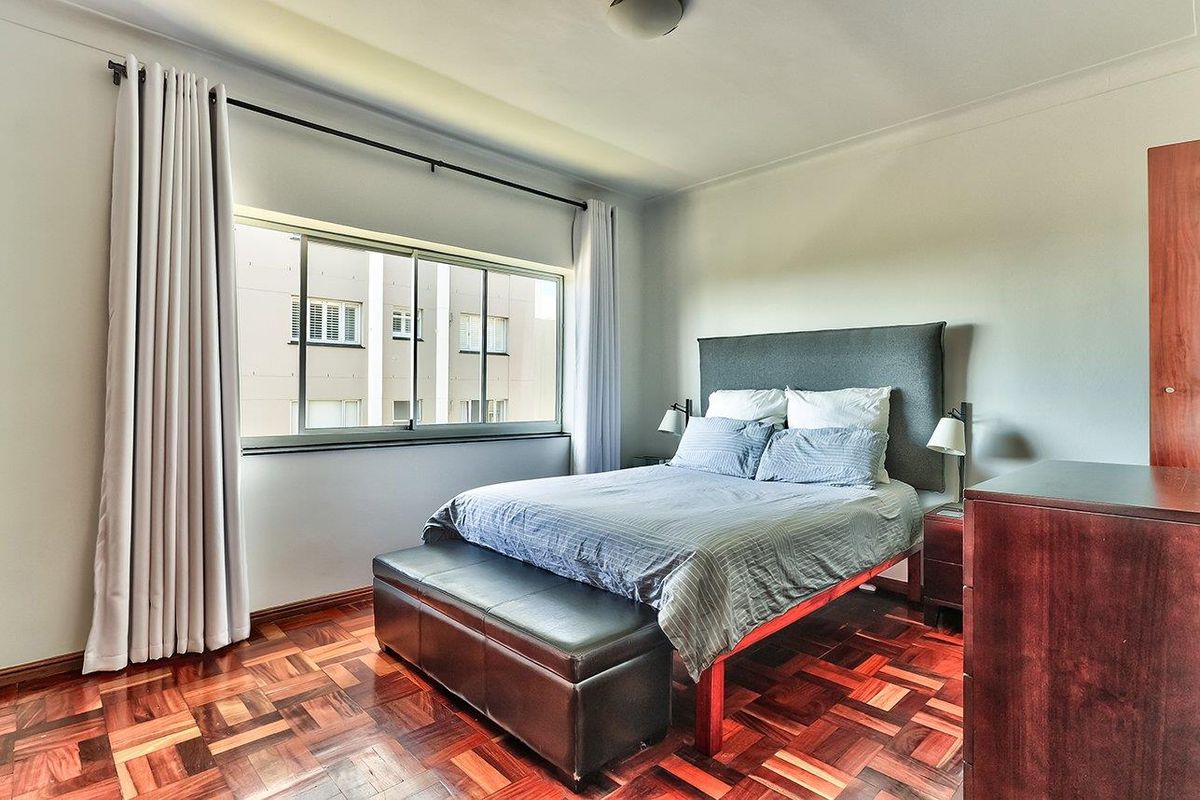A well-maintained bedroom featuring classic parquet flooring, a large sliding window for natural light, and neutral wall finishes.