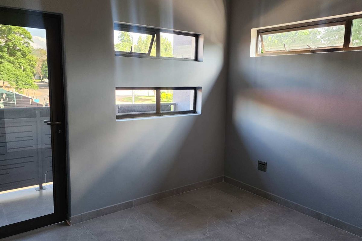 A bright, empty room featuring modern grey tiled flooring, dark-framed windows, and a glass-panelled door leading to an outdoor area.