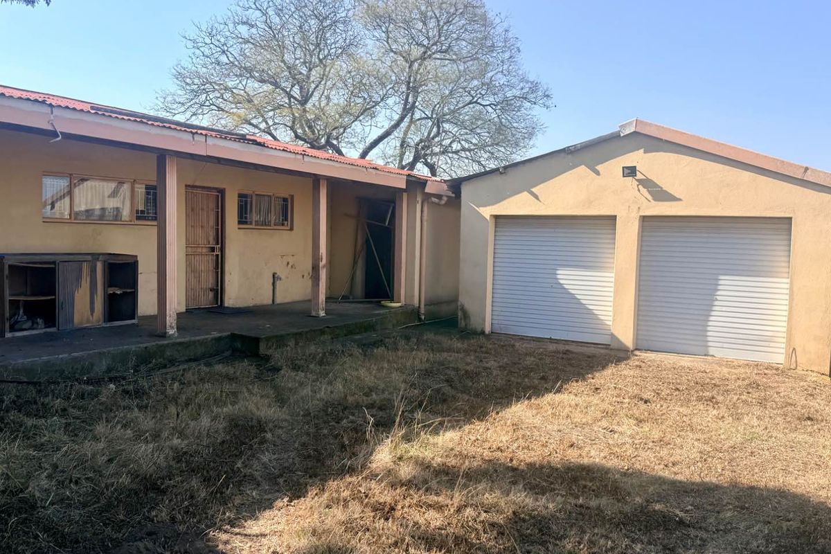 Rear view of the property featuring a covered patio area, adjacent double garage with roller doors, and an expansive, unmaintained garden space.