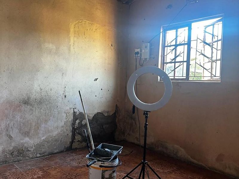An empty bedroom space featuring tiled flooring, security bars on the window, and exposed corrugated roof sheeting, currently undergoing wall repairs.