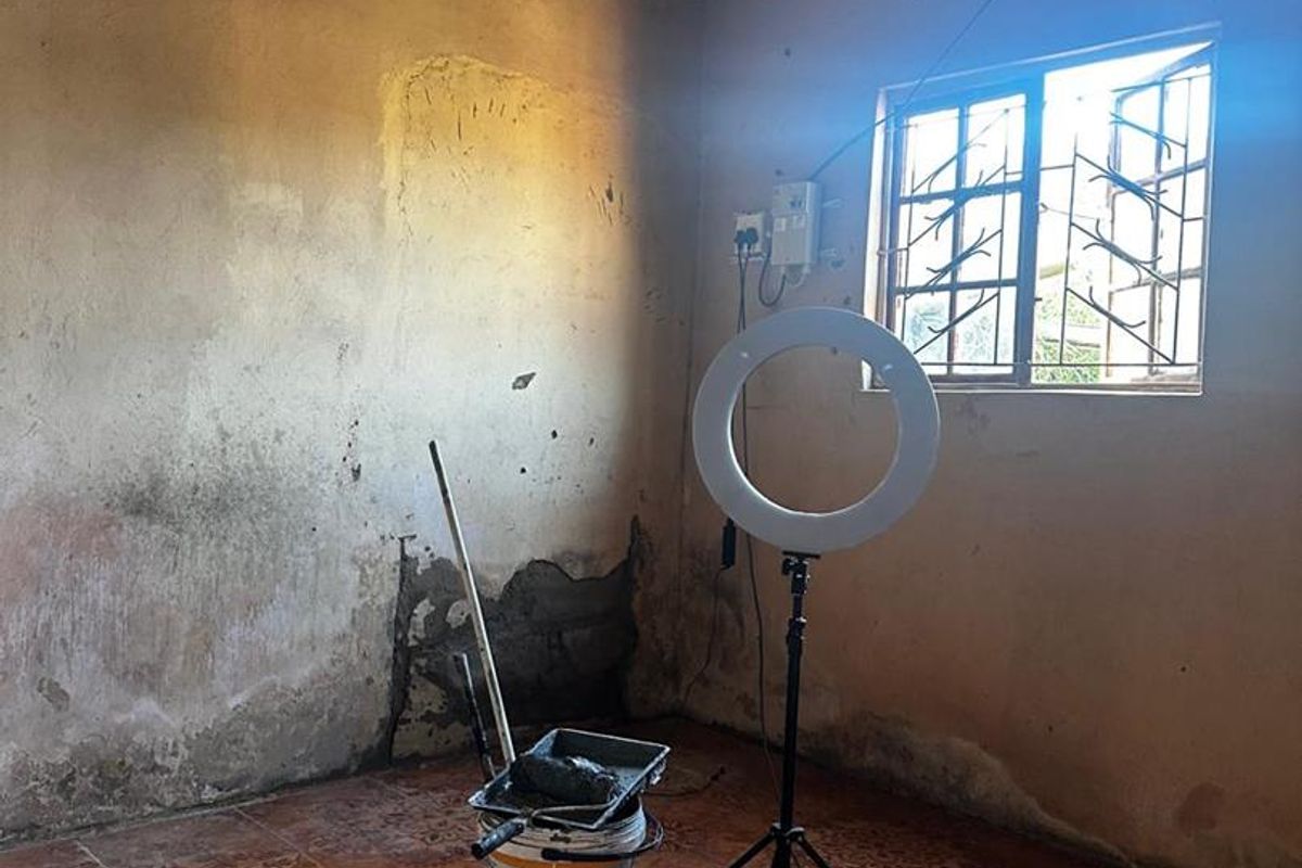 An empty bedroom space featuring tiled flooring, security bars on the window, and exposed corrugated roof sheeting, currently undergoing wall repairs.