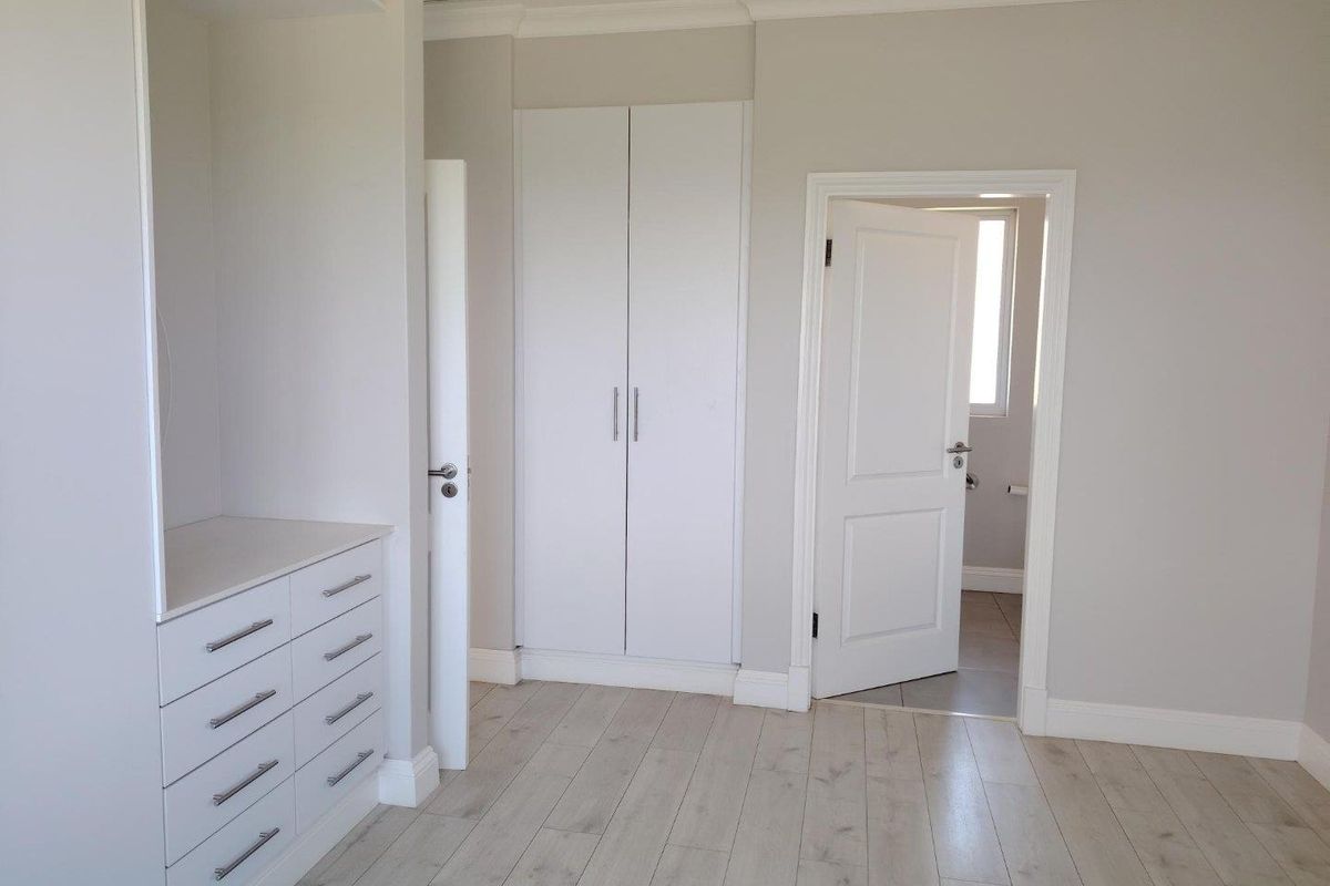 A bright, modern bedroom featuring neutral walls, light laminate flooring, and ample built-in cupboard storage, with an adjoining doorway leading to a bathroom.
